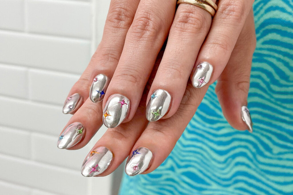 duas mÃ£os com unhas prateadas, com efeito cromado e pedras coloridas