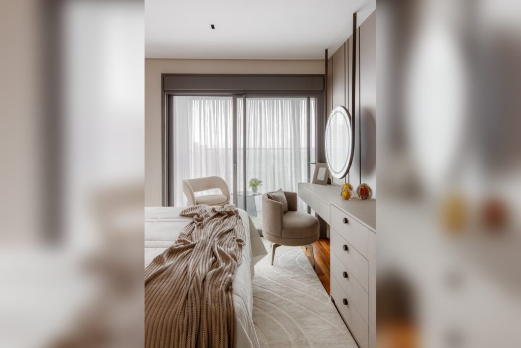 Quarto com cama coberta por manta de tricÃ´ bege, tapete claro e poltrona prÃ³xima Ã  escrivaninha com espelho redondo iluminado
