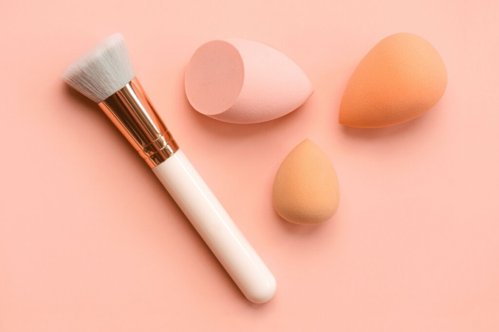 Pincel branco ao lado de trÃªs esponjas de maquiagem e sobre um fundo rosa-claro