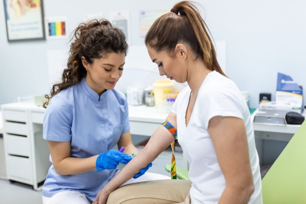 Mulher com o cabelo cacheado preso no alto da cabe&Atilde;&sect;a, usando jaleco de enfermeira, com outra mulher usando camiseta branca e cal&Atilde;&sect;a bege e com o cabelo preso em rabo de cavalo em consult&Atilde;&sup3;rio m&Atilde;&copy;dico, enfermeira est&Atilde;&iexcl; preparando a paciente para doa&Atilde;&sect;&Atilde;&pound;o de sangue