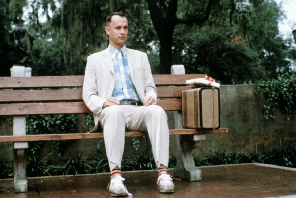 Forrest Gump, interpretado por Tom Hanks, sentado em um banco com uma mala ao lado em cena do filme
