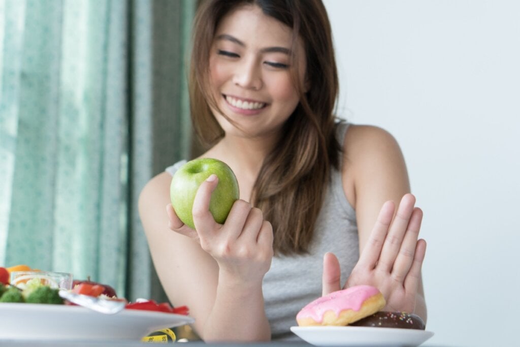 Jovem com cabelo liso, solto e usando uma regata cinza com uma maçã verde na mão e com a outra recusando um donut