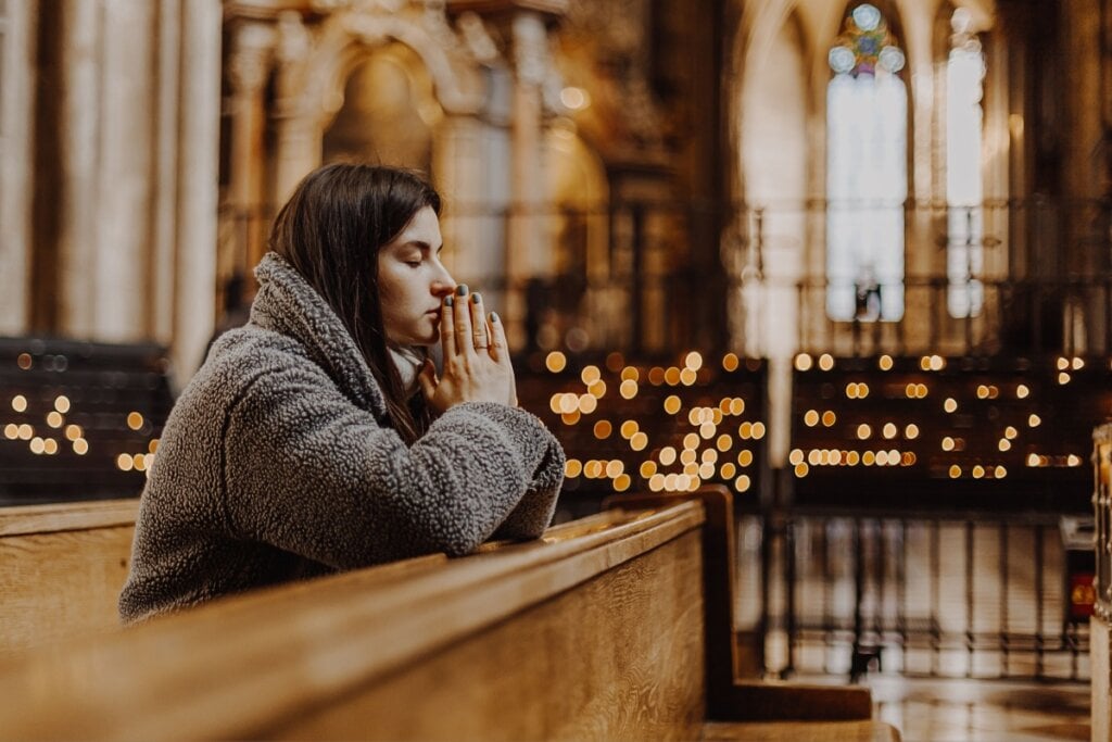 Jovem usando roupas de frio cinza e o cabelo solto, rezando ajoelhada na igreja de olhos fechados