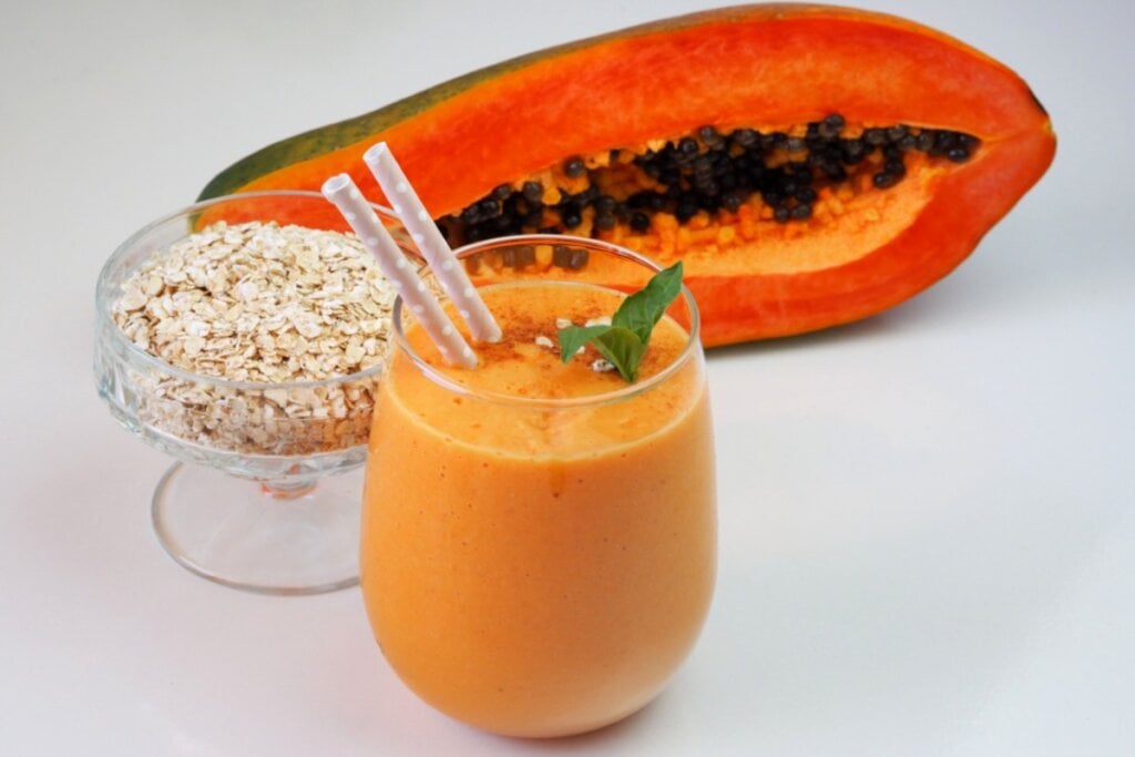 Copo de vitamina de mamÃ£o e aveia, aveia em grÃ£os em recipiente ao lado e mamÃ£o cortado ao meio ao fundo