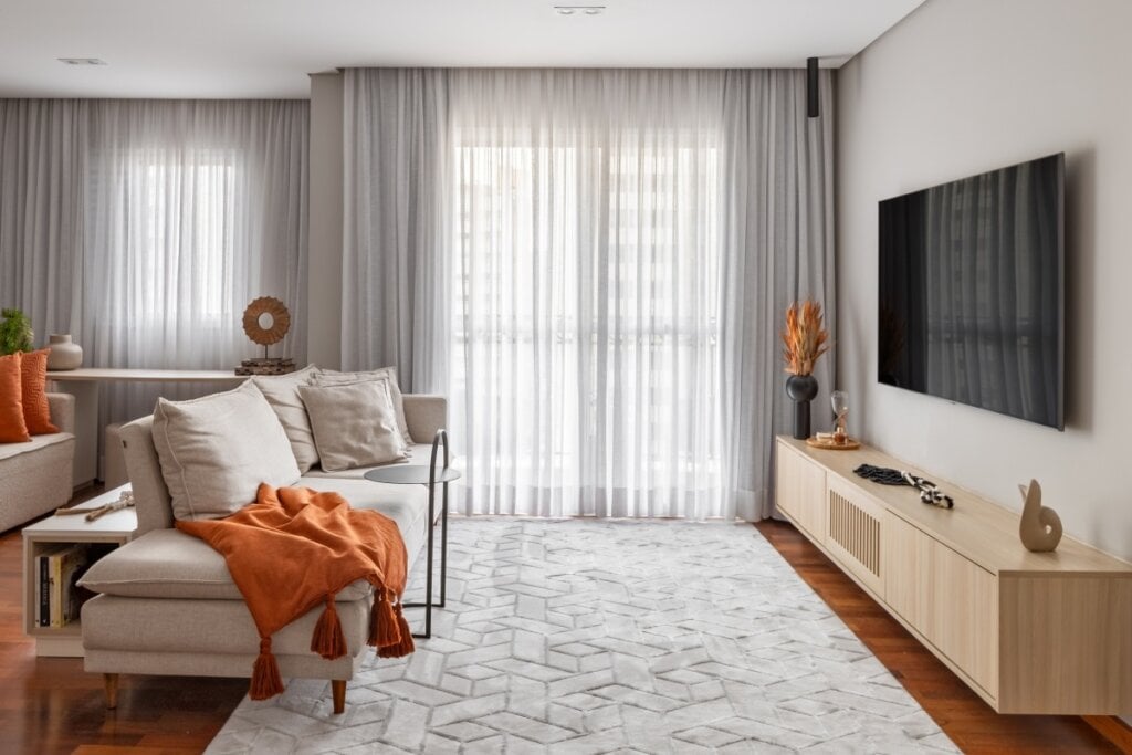 Sala de estar com sofÃ¡ claro decorado com almofadas e manta laranja, cortinas translÃºcidas e tapete geomÃ©trico sobre piso de madeira