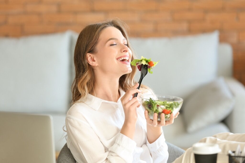 Mulher segurando pote de salada e levando garfo com salada Ã¡ boca