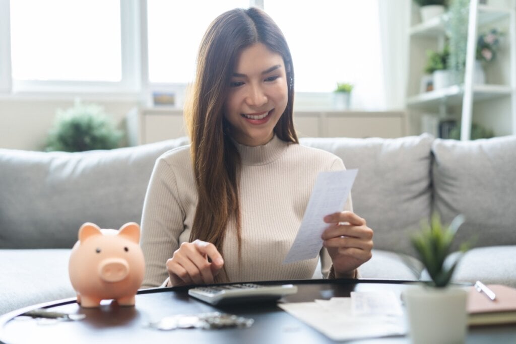 mulher de blusa bege, sorrindo, segurando papel e digitando na calculadora em cima da mesa com cofrinho de porco rosa ao lado