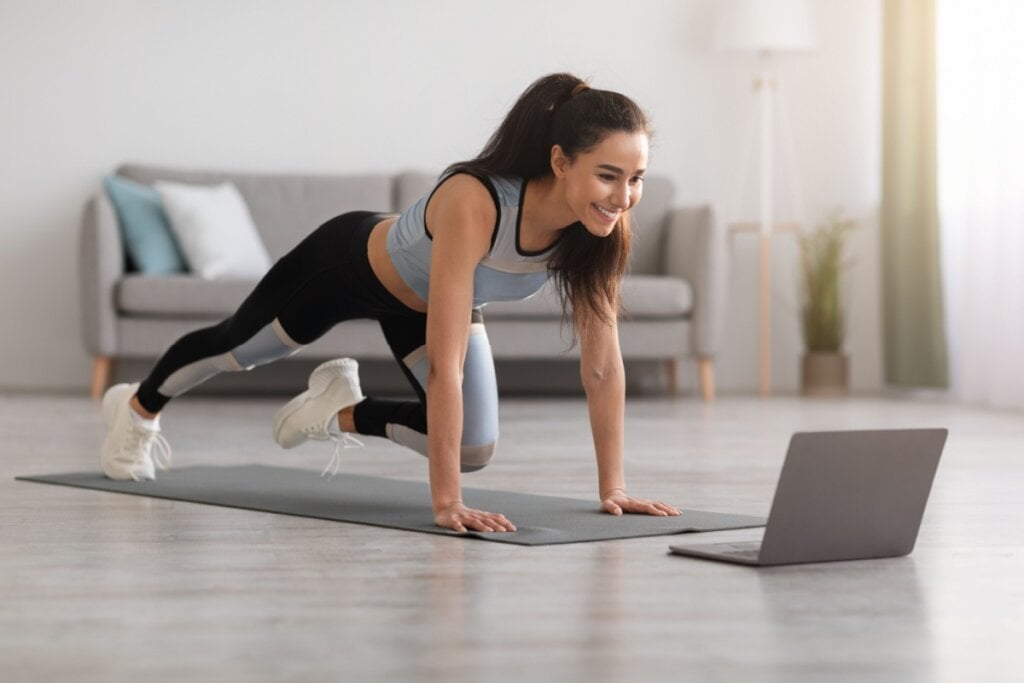 Mulher com roupa de academia praticando exercÃcio na sala de casa, assistindo a um vÃdeo no notebook