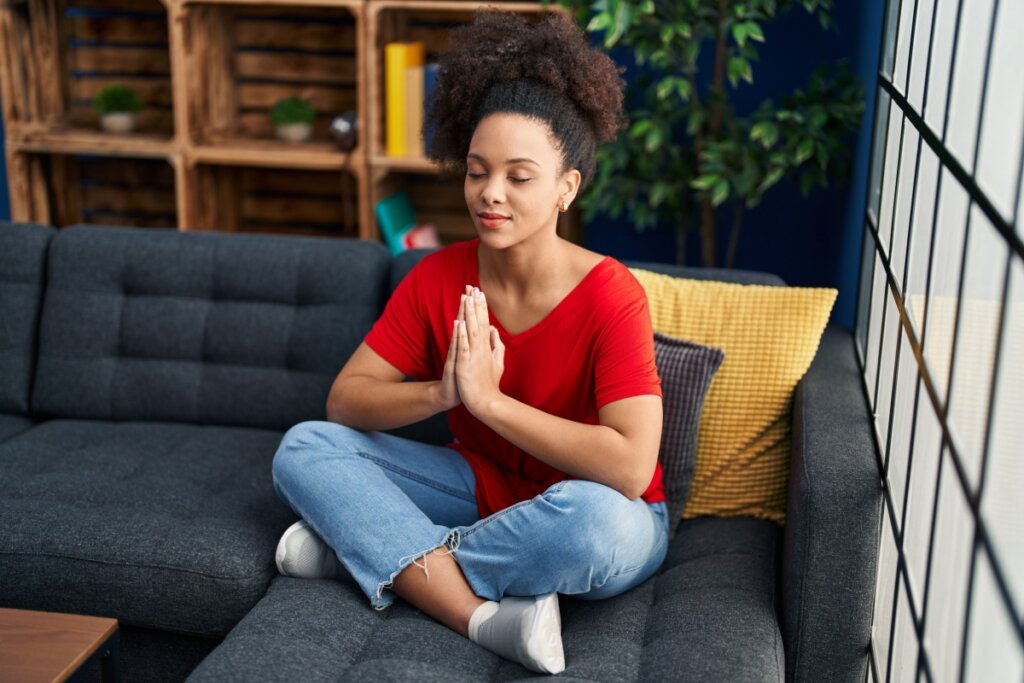 Mulher sentada em posi&Atilde;&sect;&Atilde;&pound;o de medita&Atilde;&sect;&Atilde;&pound;o sobre o sof&Atilde;&iexcl;, com olhos fechados e m&Atilde;&pound;os em prece