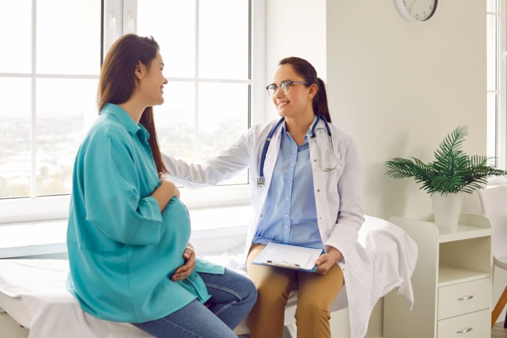 Paciente grÃ¡vida com o cabelo liso, solto e usando uma camisa verde ao lado da mÃ©dica que estÃ¡ com o cabelo preso, usando Ã³culos e camisa azul e um jaleco branco