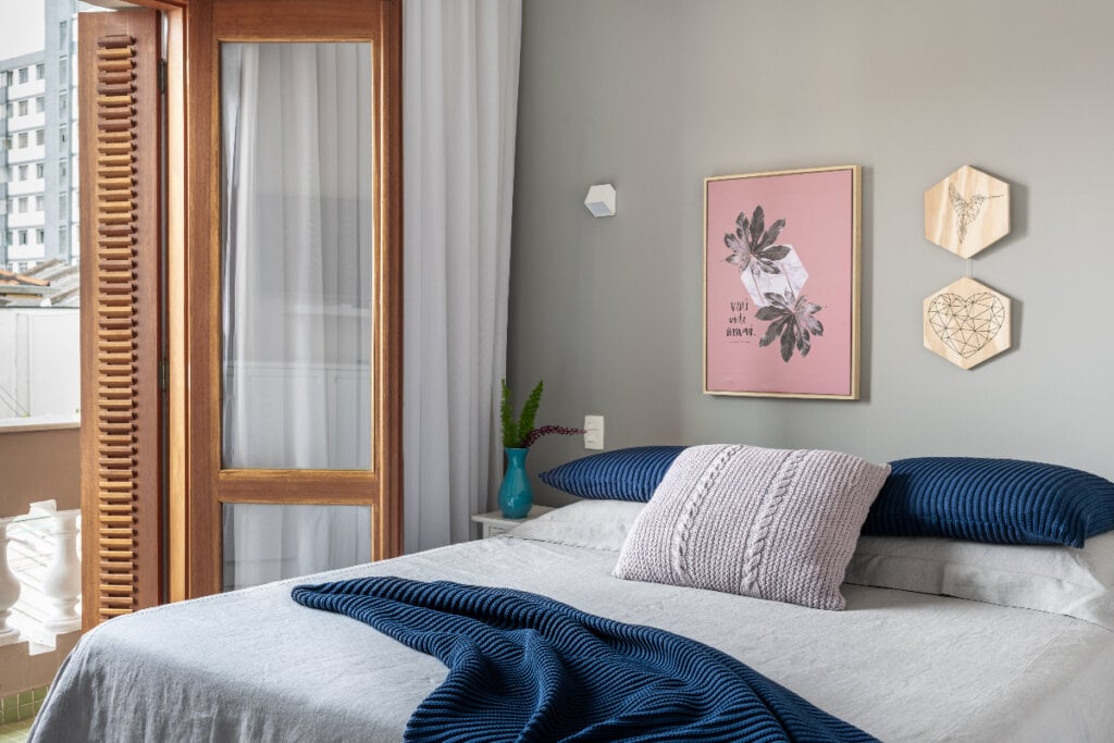 quarto com cama cinza, cobertos azul, porta de madeira do lado esquerdo com cortina branca e parede cinza com quadro rosa ao fundo da cama