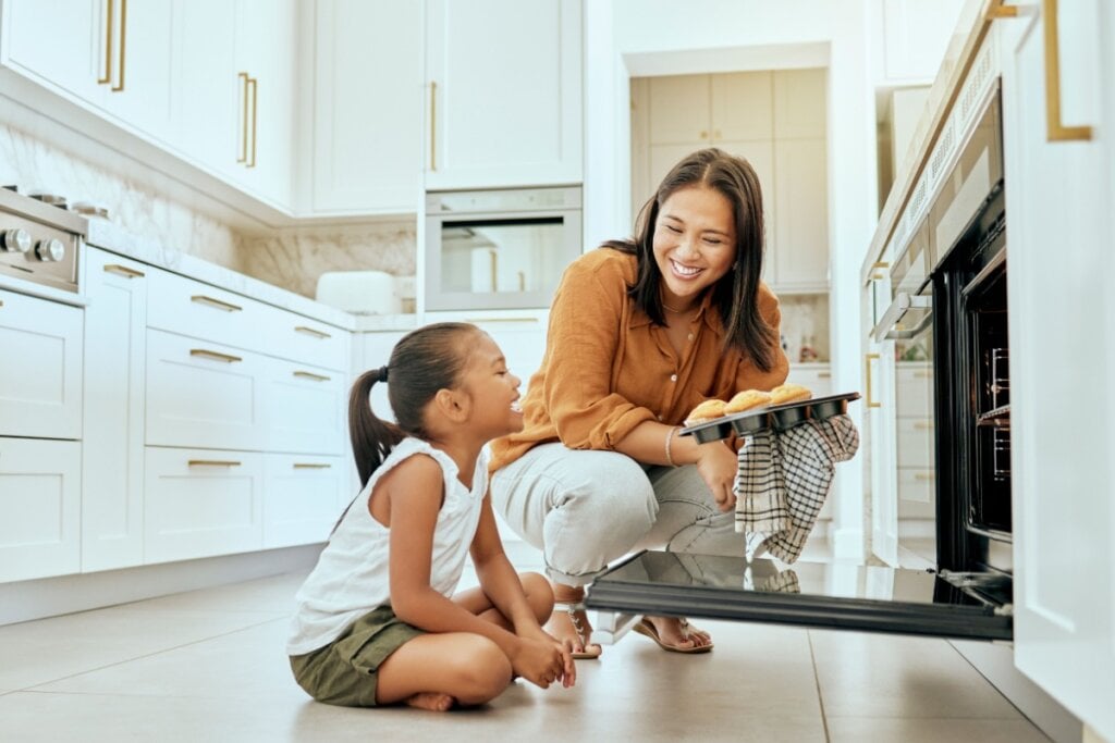 Mulher colocando uma forma com biscoitos no forno e uma menina sentada do lado olhando