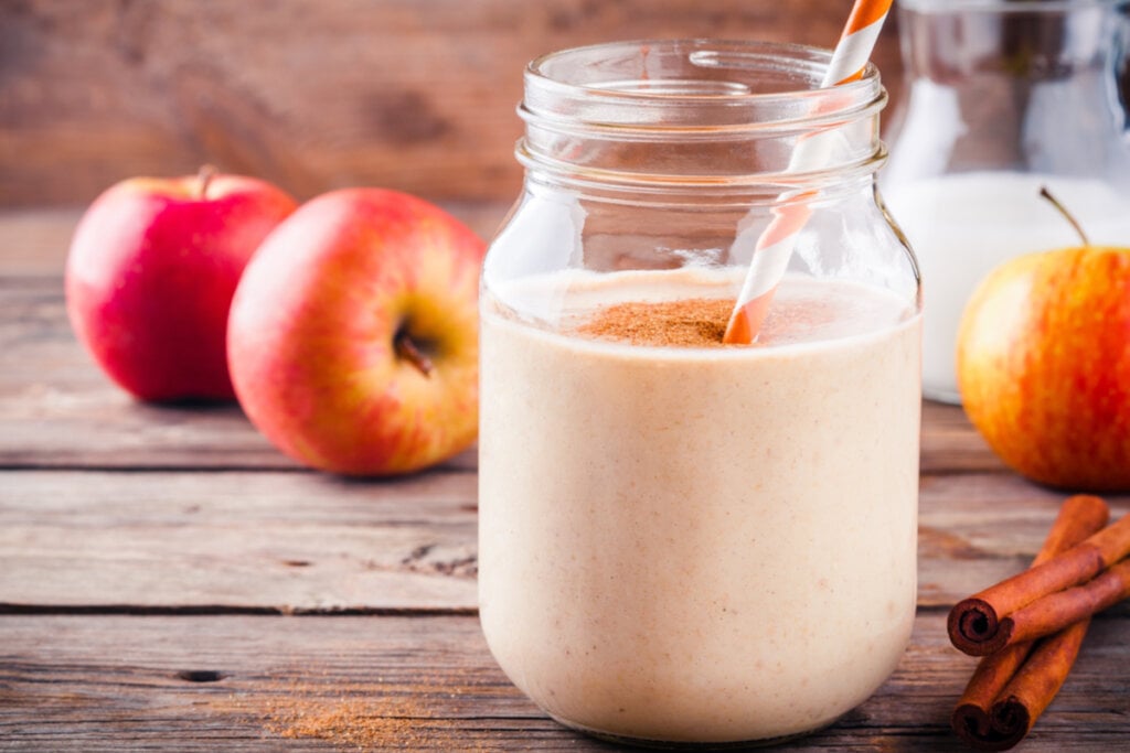 Copo com vitamina de maÃ§Ã£ e canela em pÃ³ por cima. Ao fundo, hÃ¡ maÃ§Ã£ e, ao lado do copo, canela em pau