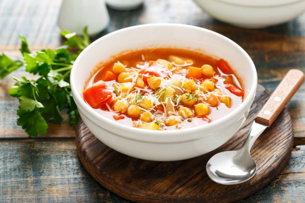 Sopa de grão-de-bico com legumes e parmesão em bowl branco ao lado de colher e folhas verdes
