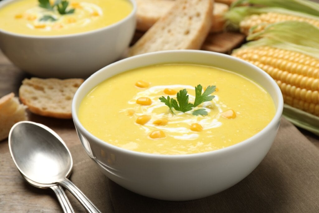 Sopa de milho em uma tigela branca e ao lado pedaços de pães e talheres