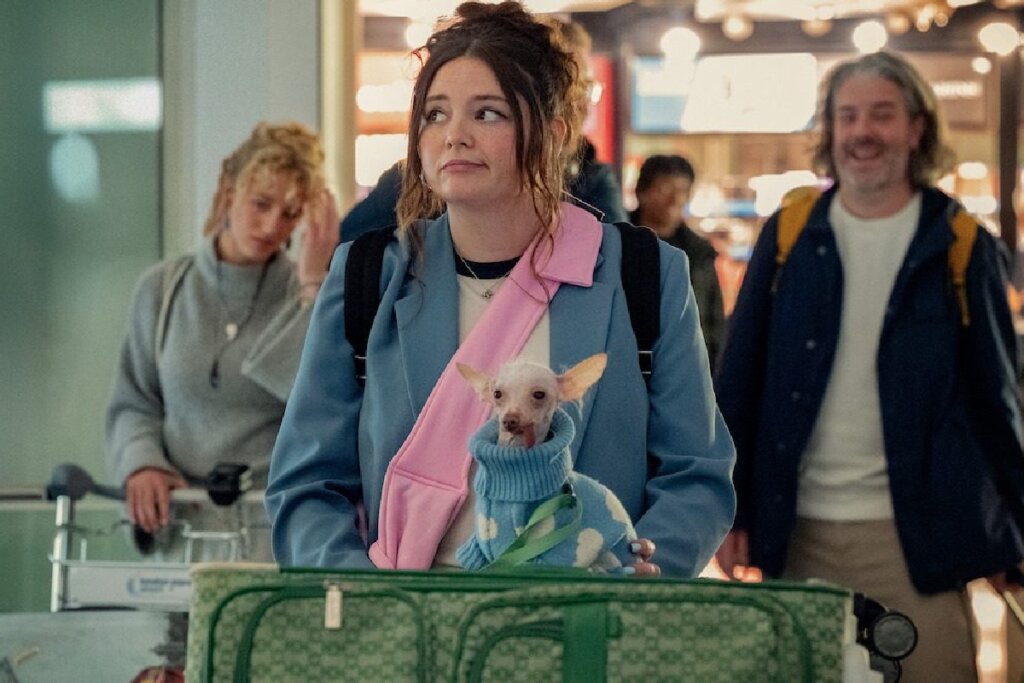 Jovem com casaco azul e faixa rosa com um cachorro pequeno de suéter em aeroporto movimentado