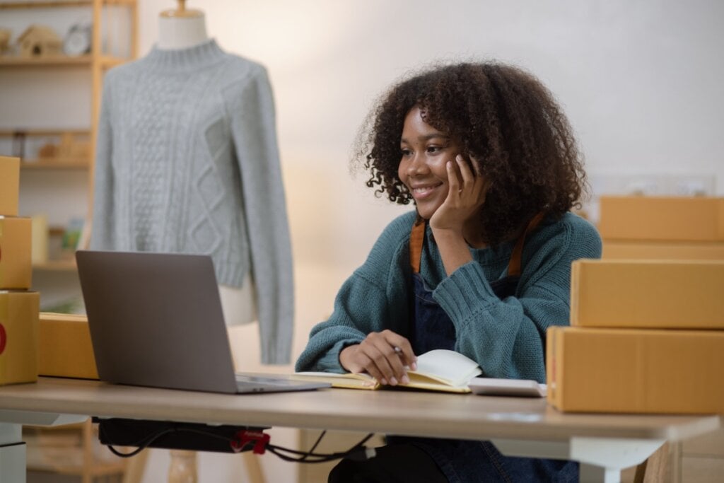 Mulher com cabelo cacheado curto usando um suÃ©ter verde e avental olhando pro computador e sorrindo.