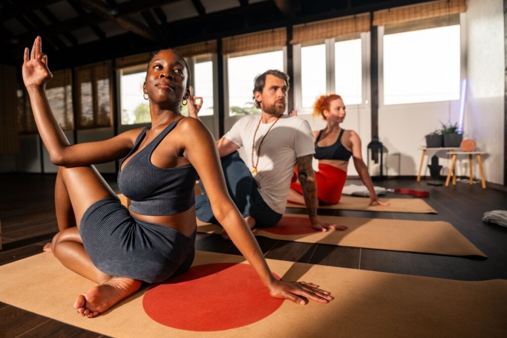duas mulheres e um homem sentados em tapete de yoga no chão e fazendo postura de torção das costas