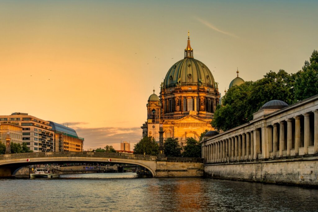 Imagem de uma construÃ§Ã£o neoclÃ¡ssica em frente a uma ponte e um rio abaixo dela ao pÃ´r do sol em Berlim