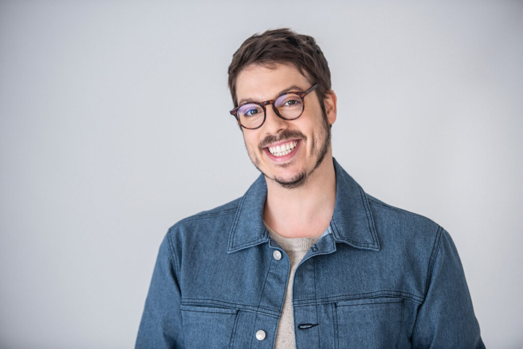 Homem com cabelo curto e barba, usando Ã³culos preto e camisa jeans em fundo claro