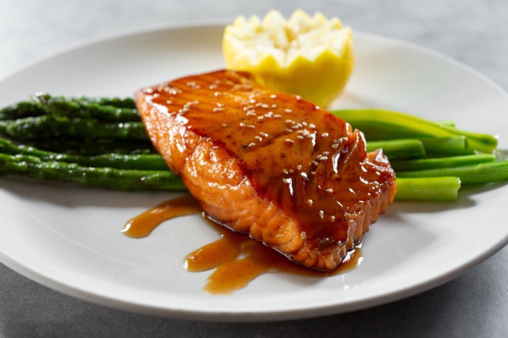 Em prato branco, salmÃ£o grelhado com molho teriyaki, aspargos e limÃ£o para decorar