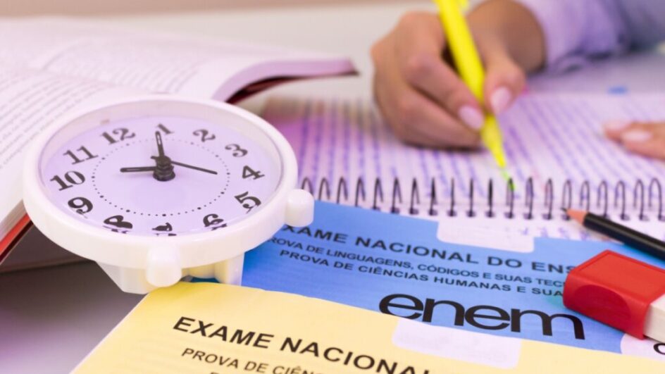 Relógio branco em cima de provas do Enem e uma mão com marca texto em cima de um caderno do lado