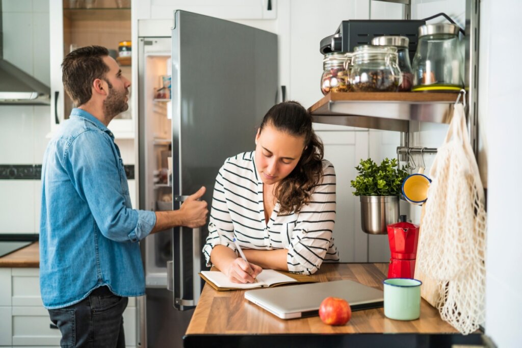 Casal na cozinha montando lista de compras, o homem estÃ¡ usando uma camisa jeans e calÃ§a preta e conferindo os alimentos na geladeira e a mulher estÃ¡ fazendo a lista de compras, ela estÃ¡ com o cabelo preso e usando uma camisa de manga longa listrada