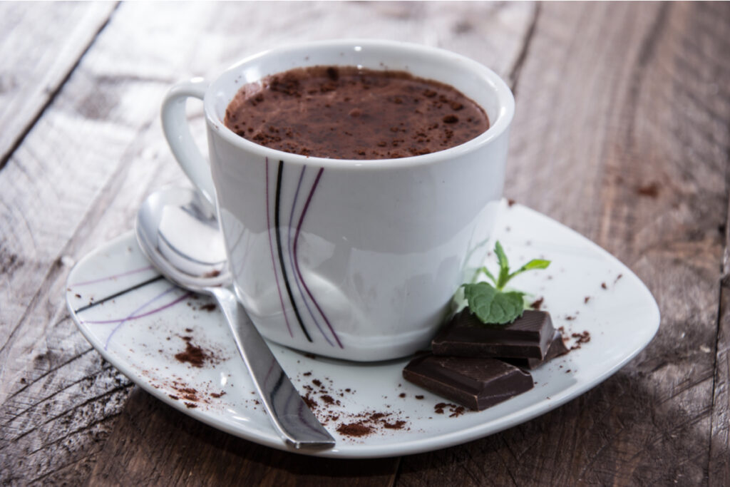 xícara branca em pires branco com chocolate quente com cacau em pó. dois tabletes de chocolate com hortelã no pires