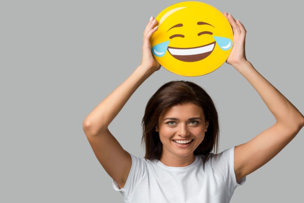 Mulher sorridente segurando acima da cabeÃ§a um grande emoji amarelo de rosto rindo com lÃ¡grimas, sobre fundo cinza