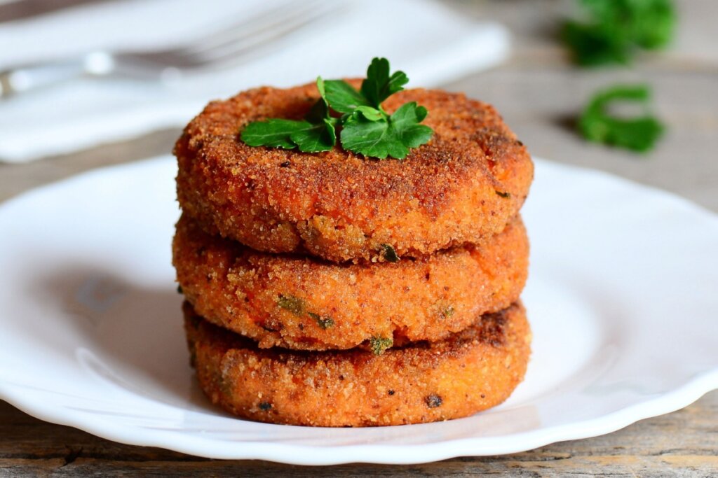 HambÃºrguer de cenoura empanado servido em prato de cerÃ¢mica branco em cima de mesa de madeira