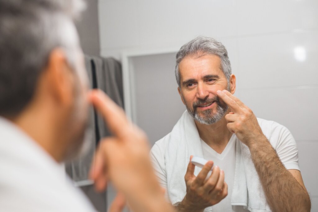 Homem com cabelo e barba grisalhos, usando camiseta branca, com toalha no ombro em frente ao espelho aplicando produto protetor na barba