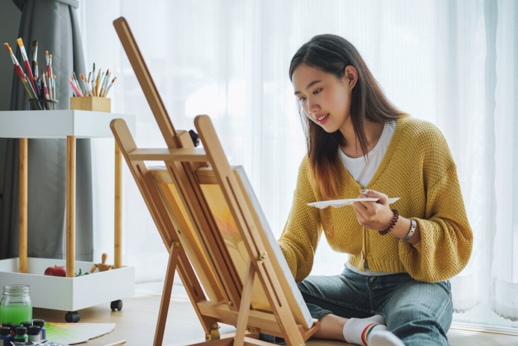 Jovem com o cabelo liso, escuro e solto. Usando uma camiseta branca e um suÃ©ter amarelo, calÃ§a jeans e uma meia branca com detalhes coloridos, sentada no chÃ£o pintando tela em cavalete
