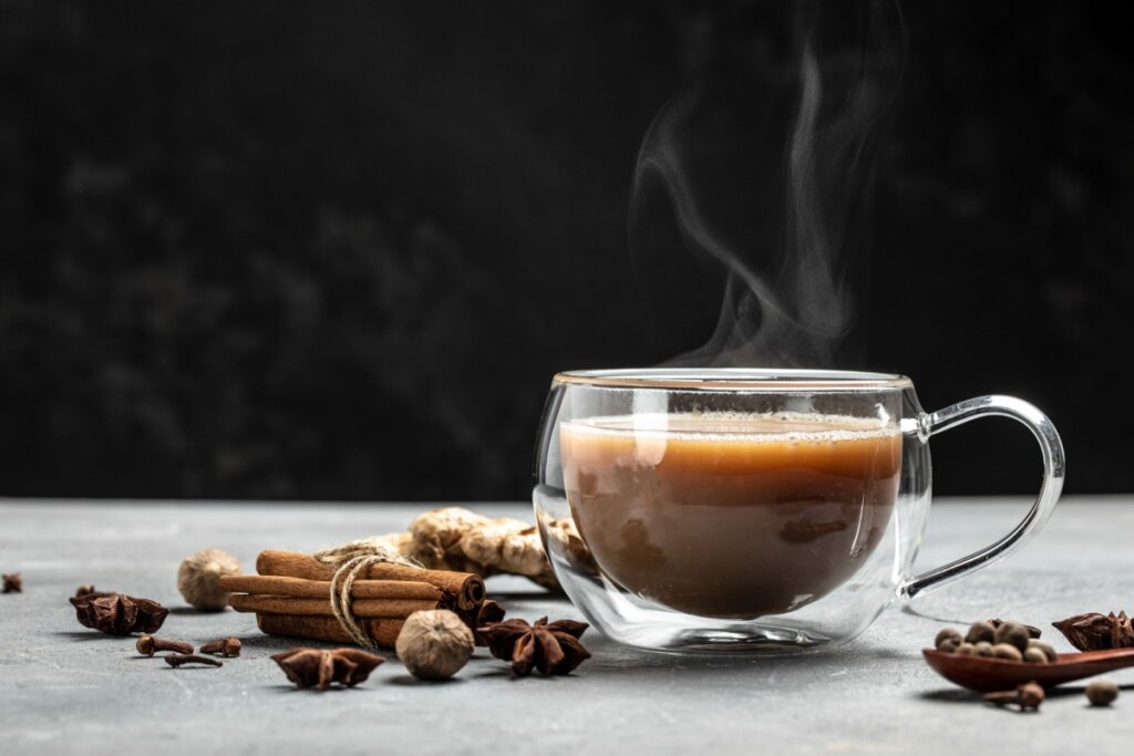 Masala chai servido em xÃcara de vidro em cima de mesa de madeira com os ingredientes ao redor da xÃcara como canela, cravos da Ãndia