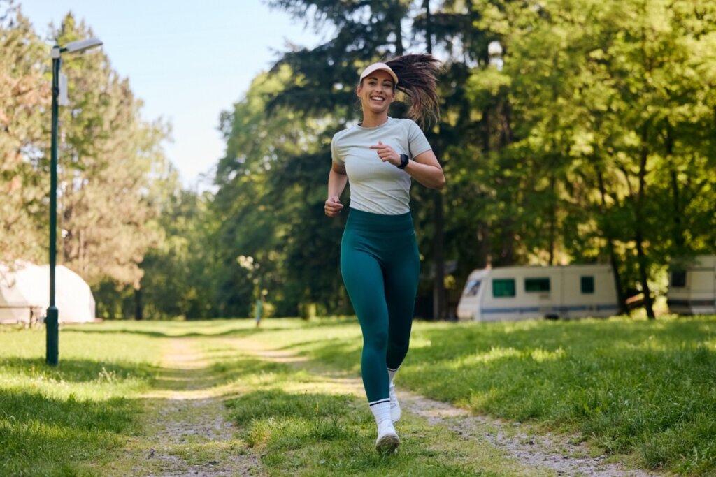 Mulher com cabelo amarrado, usando viseira, uma camiseta cinza e uma calça verde correndo no parque
