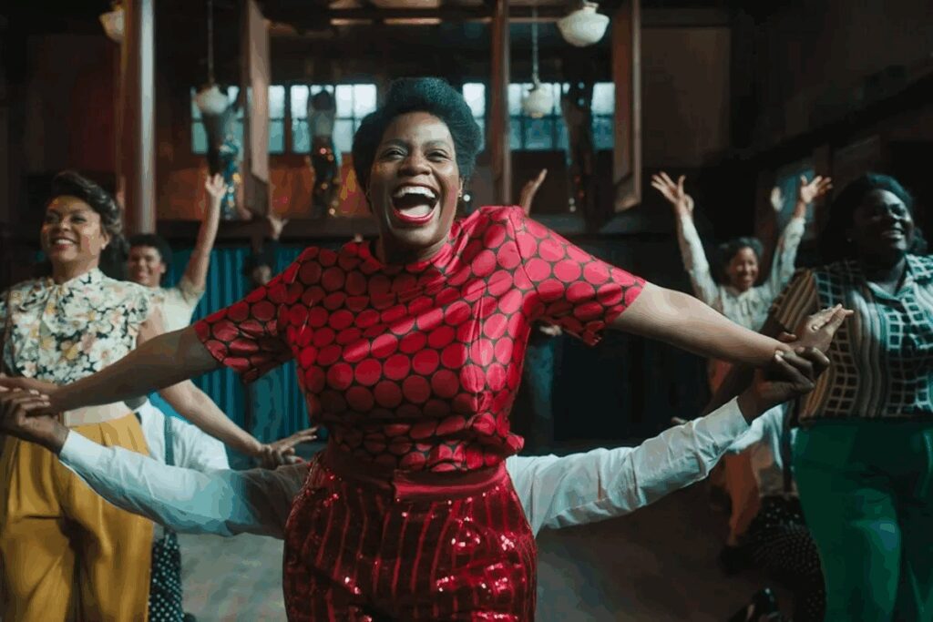 Mulheres com roupa de época dançando, uma mulher negra usando um vestido vermelho está no centro da imagem de braços abertos e sorrindo