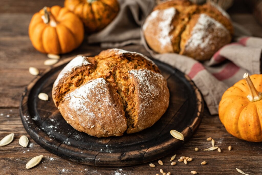 PÃ£o doce rÃºstico de abÃ³bora polvilhado com farinha, rodeado por sementes e pequenas abÃ³boras