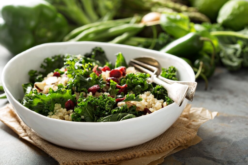 prato branco em cima de pano marrom com salada de couve com quinoa, cranberries e amÃªndoas. alimentos verdes desfocados ao fundo