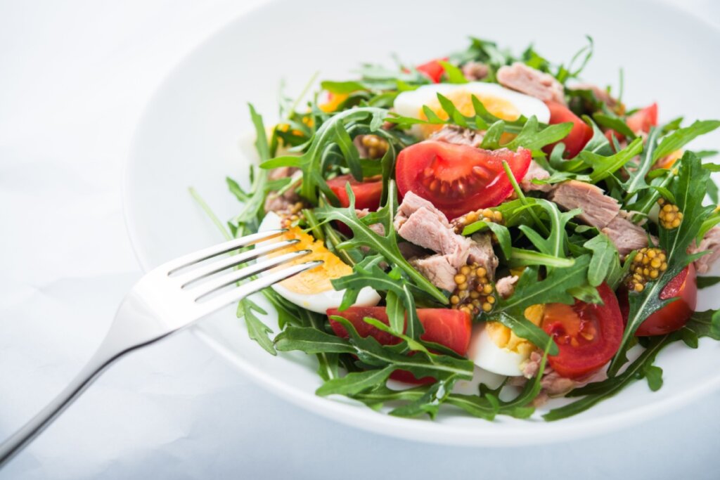 Salada de atum com ovo, rÃºcula e tomate servido em prato branco em cima de mesa branca com garfo ao lado
