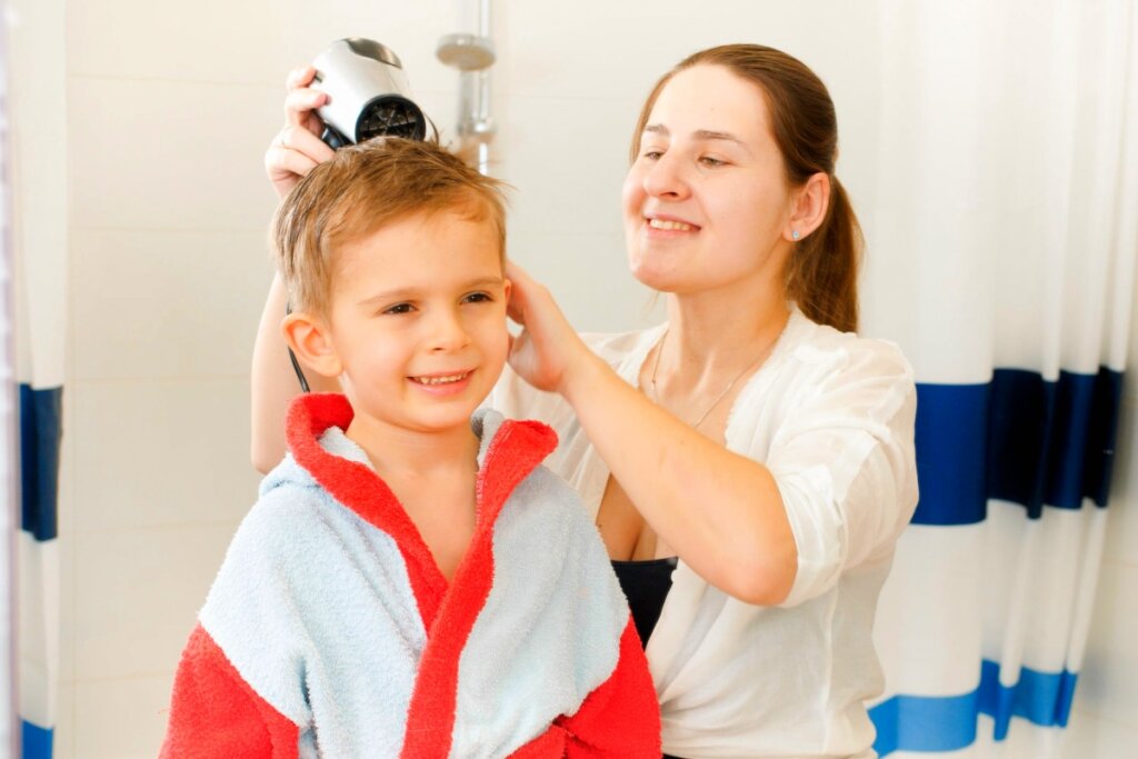 MÃ£e secando o cabelo do filho com secador apÃ³s atividade aquÃ¡tica, enquanto o menino sorri usando um roupÃ£o de banho