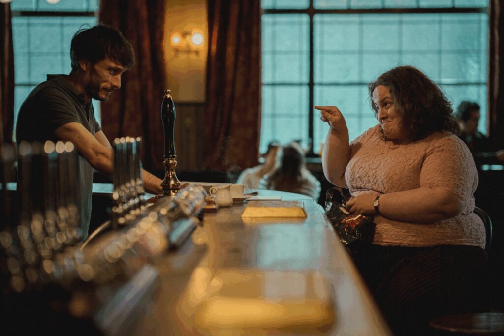 Homem com cabelo curto e barba, usando camiseta polo escura servindo uma bebida a uma mulher com o cabelo cacheado e usando camiseta estampada com uma bolsa na mÃ£o enquanto aponta o dedo para o homem