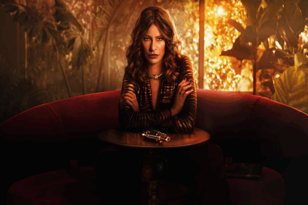 Mulher com cabelo cacheado castanho, usando blusa de manga longa estampada, sentada em sofÃ¡ curvo vermelho na frente de uma de centro com uma arma ao fundo uma floresta pegando fogo