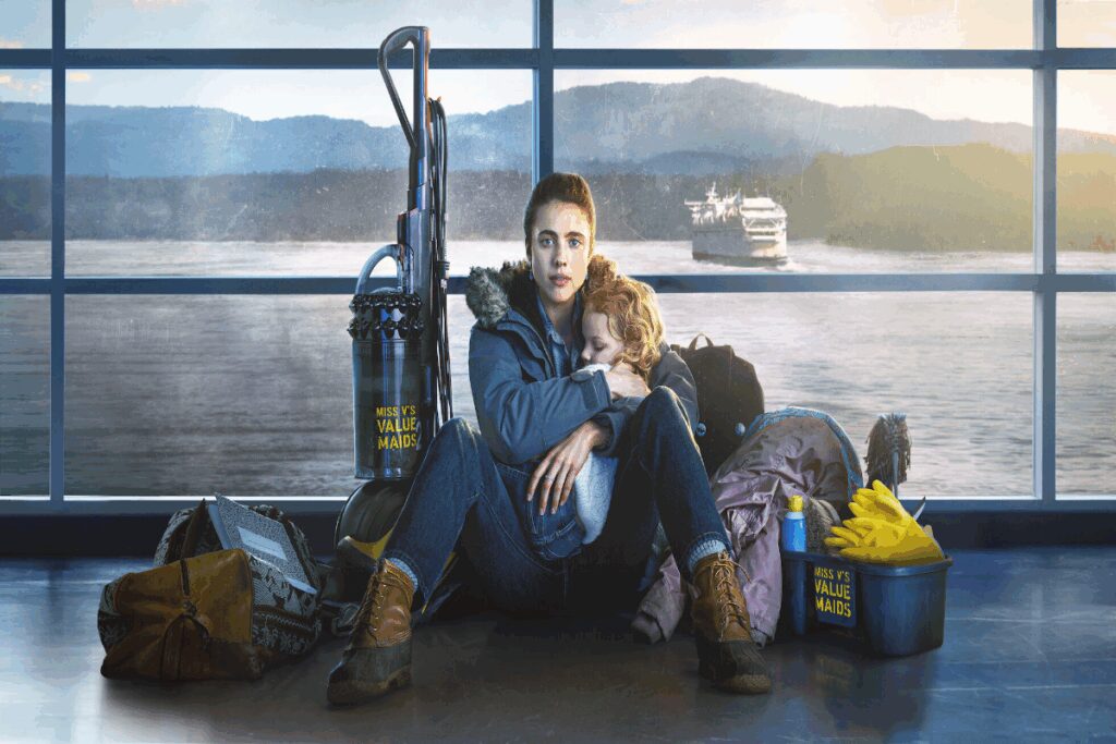 Mulher com roupas de frio sentada no chÃ£o com crianÃ§a no colo ao redor dela estÃ£o itens de limpeza e atrÃ¡s uma parede de vidro