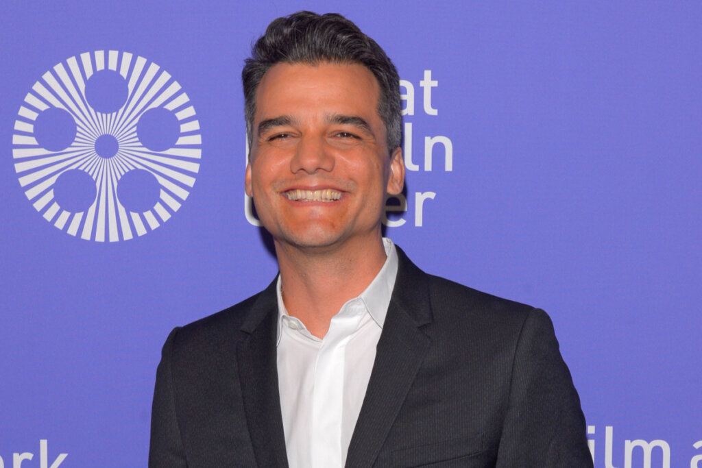 Homem com cabelo preto curto, usando camisa branca e terno preto sorrindo posando para foto em fundo roxo