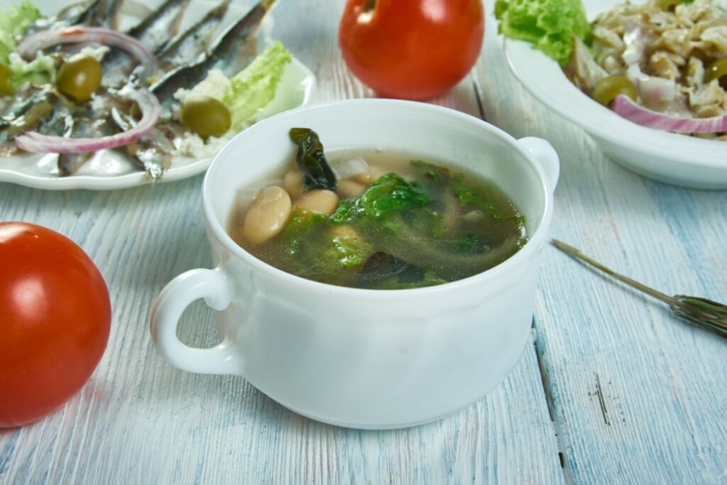 Mini panela branca com sopa de escarola com feijÃ£o-branco dentro em cima de uma mesa de madeira branca