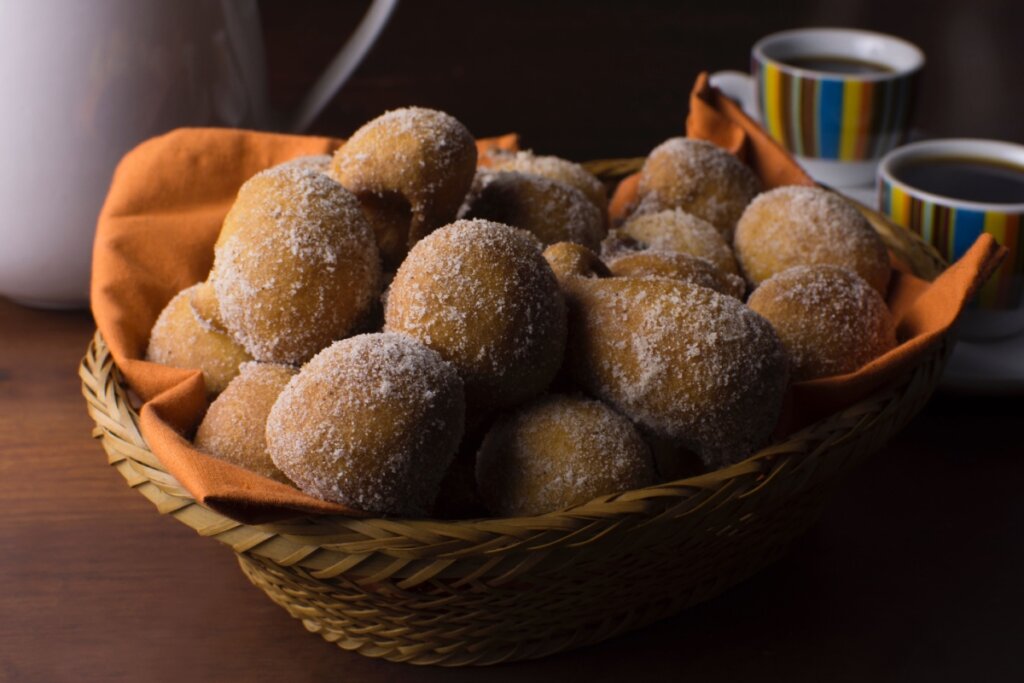 Cesta de vime repleta de bolinhos de chuva dourados e açucarados, acompanhada de xÃcaras de café