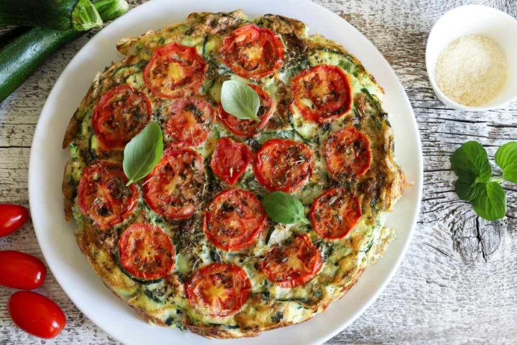 Prato branco com fritada de abobrinha e tomate decorada com folhas de manjericÃ£o