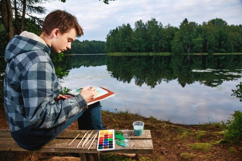 Adolescente sentado Ã  beira de um lago pintando com aquarela em um momento de introspecÃ§Ã£o