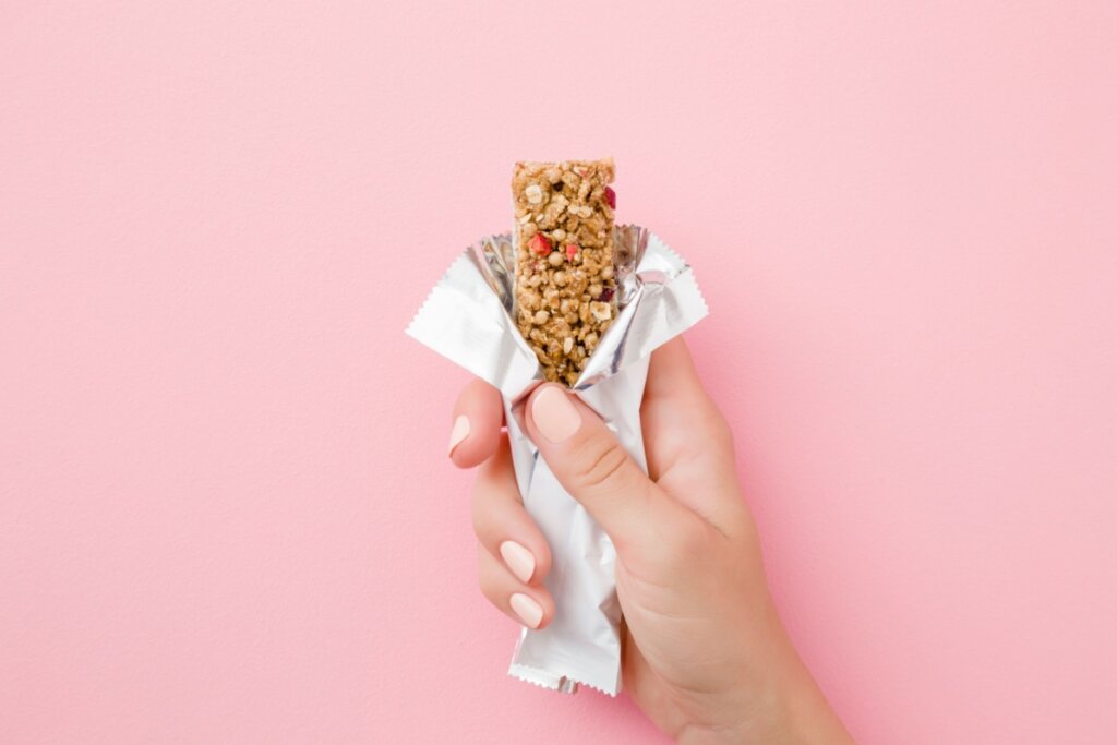 Uma mão segurando uma barrinha de cereal com a embalagem aberta em fundo rosa