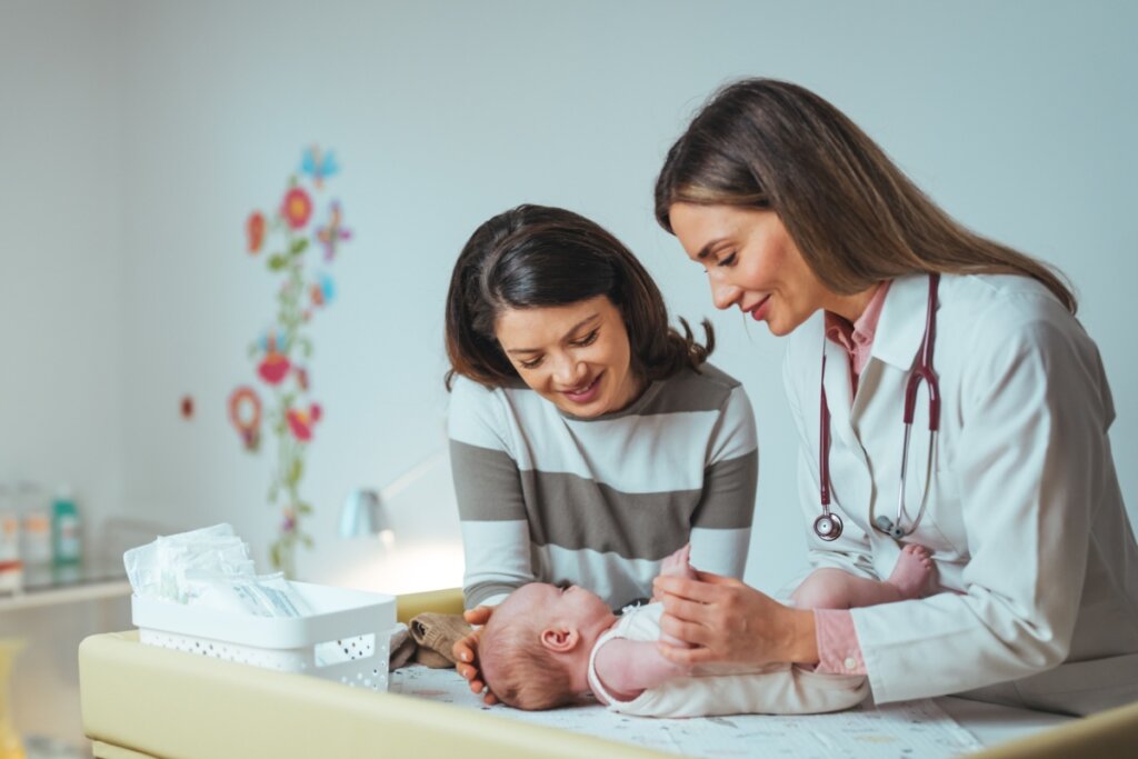 BebÃª em consulta com pediatra e com a mÃ£e ao lado. Uma das mulheres tem o cabelo curto e liso, e estÃ¡ usando uma camisa de manga longa listrada e a outra tem o cabelo longo e liso, e estÃ¡ usando uma camisa rosa de botÃµes e um jaleco branco com um estetoscÃ³pio no pescoÃ§o