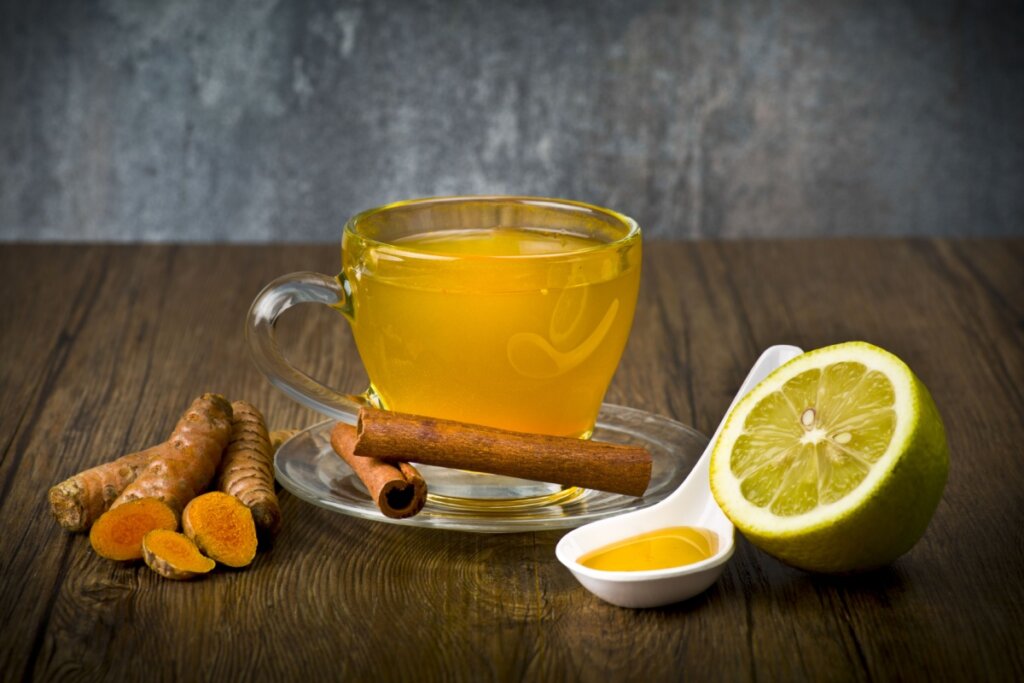 ChÃ¡ de cÃºrcuma com canela e limÃ£o servido em xÃ­cara de vidro em cima de mesa de madeira com mel, limÃ£o e cÃºrcuma ao lado